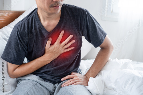 Young Asian Man Sitting on Bed, Clutching His Chest in Discomfort and Pain A of Heart Disease Symptoms Including Angina, Cardiovascular Problems, and Hypertensive Effects.
