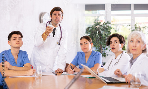 Middle-aged male hospital chief turning on presentantion for young adult mature doctors and nurses sitting with reports and laptops at table