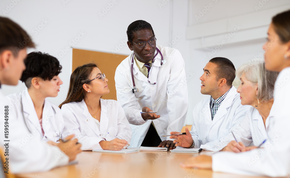 Group of medical students in white coats studying in classroom. African-american man professor talking to medical interns. Group of medical students in white coats studying in classroom. African-american man professor talking to medical interns.