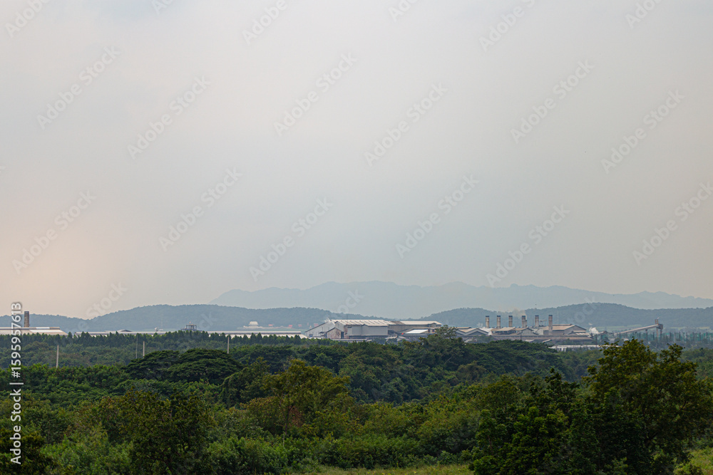 Obraz premium View of industrial factory complex surrounded by green forest with distant mountains under cloudy sky, showing contrast of nature and industry.