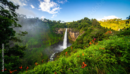 Tall waterfall flowing between green cliffs and dense tropical forest with refreshing mist.