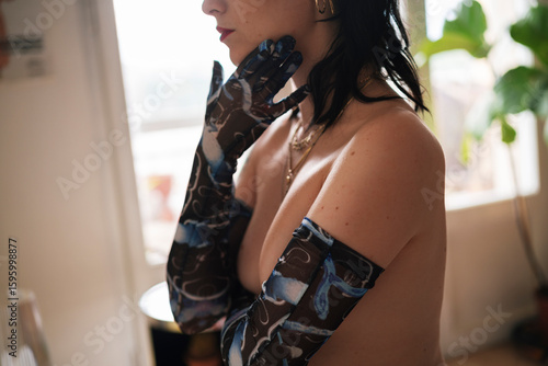 Side view crop topless slim female wearing long gloves covering breast and touching chin while standing in light room