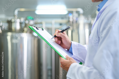 Scientist in lab coat writing on clipboard in laboratory with large tanks in the background
