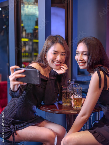 Two young women taking a selfie together at a bar while enjoying drinks. They are smiling happily, dressed stylishly, and sitting at a round table with beer glasses