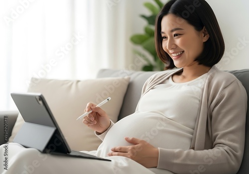 Joyful Pregnant Asian Woman Sits Comfortably On A Couch, Gently Holding Her Belly While Using A Tablet And Stylus, Maternity Leave, Online Preparation