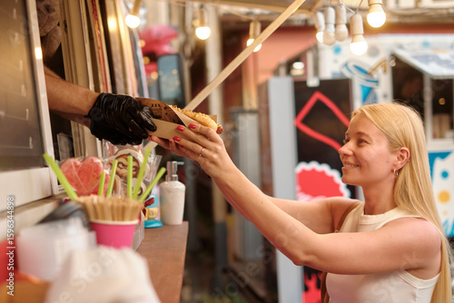 Joyful young blonde woman exchanges cash for food at lively outdoor market food truck with string stringlights, warm glow creating friendly, festive atmosphere