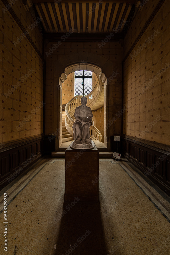 Naklejka premium Historic statue illuminated by natural light in an abandoned French chateau with a spiral staircase in the background