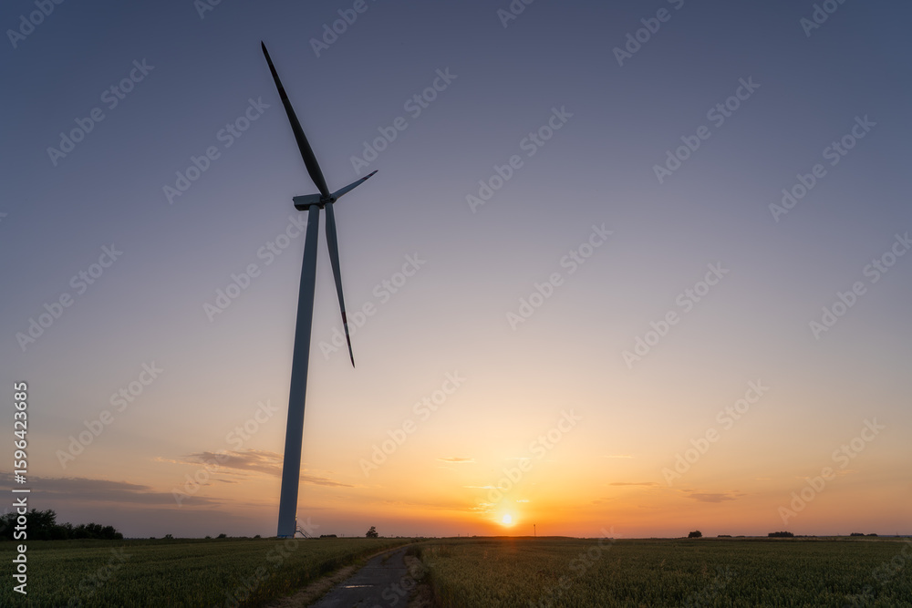 Obraz premium Wind turbine silhouette in rural field during colorful sunset – renewable energy and clean power concept with scenic countryside and environmental sustainability theme.