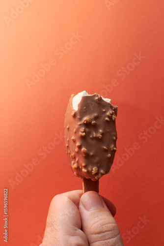 Chocolate-covered ice cream bar with nuts held in a hand, isolated on a colorful orange background.  Summer dessert, heat relief.