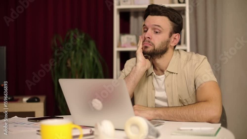 Man sitting at a desk, looking tired and frustrated while working on a laptop. His hand supports his head in exhaustion. Home office scene with warm lighting and scattered objects.