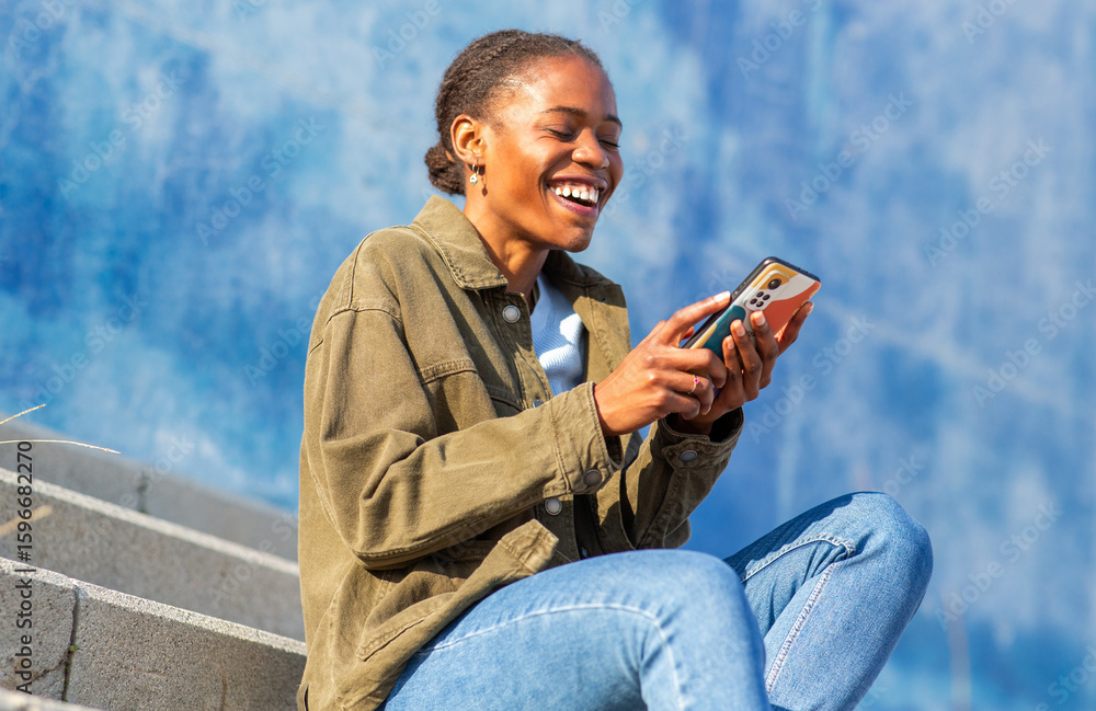 Fototapeta premium African young woman laughing while using smartphone on outdoor steps on blue background