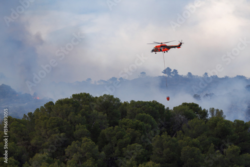 Helicoptero no reconocible con Bambi Bucket se aproxima al fuego en incendio forestal del parque natural de la Font Roja en julio de 2025, Ibi, España