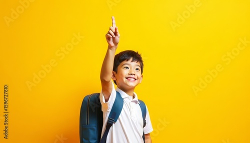 A small Asian boy in a white T-shirt smiles against a yellow background, pointing with his index finger at a place for text. I'm going to school soon. Knowledge day. The student goes to the 1st grade.