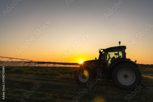 Tractor and agricultural watering pivot irrigation system on a corn field at sunset