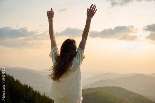 A woman stands on a mountain ridge with arms raised high, celebrating the beauty of nature as the sun sets in the background. Surrounding hills are covered in rich greenery, creating serene atmosphere