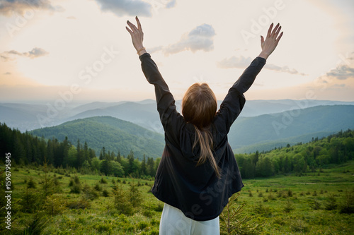 A woman stands on a mountain ridge with arms raised high, celebrating the beauty of nature as the sun sets in the background. Surrounding hills are covered in rich greenery, creating serene atmosphere