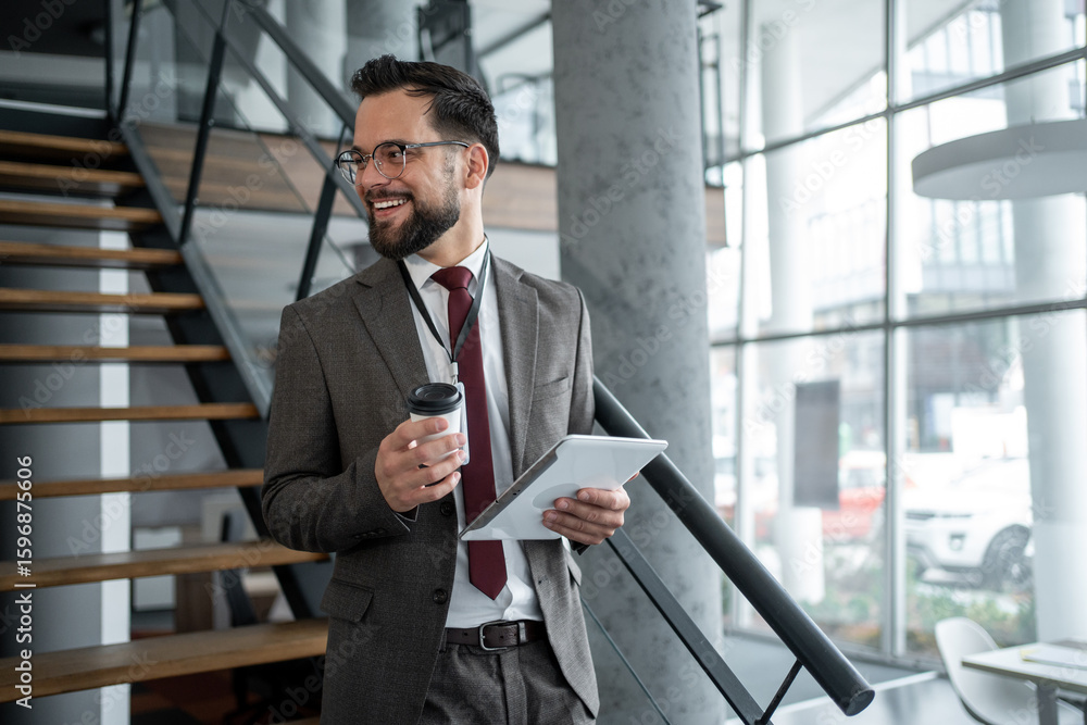Fototapeta premium Smiling businessman holding tablet and coffee, walking up stairs in office