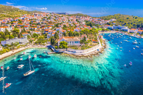 Drone image of Hvar town showing the scenic waterfront promenade, picturesque harbor with anchored boats, coastal buildings and vibrant Adriatic atmosphere in full daylight
