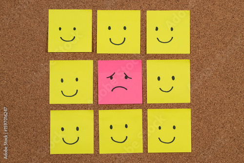 a group of sticky notes representing depression with a sad face surrounded with a lot of happy faces over a cork background