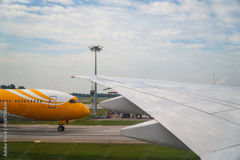 Obraz premium Aviation image capturing the wing of a commercial airplane under a cloudy sky, alongside a yellow airplane prepared on the runway. The detailed texture suggests modern aviation technology and travel.