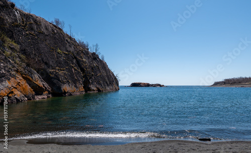 Scene looking southward at Black Beach on the North Shore of beautiful Lake Superior near Silver Bay, Minnesota, includes water, rocks, blue sky, and an island, on a sunny spring day.