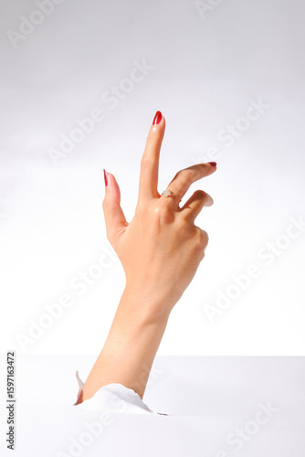 Woman’s hand with red nails and ring emerging through torn paper, pointing upward on white background