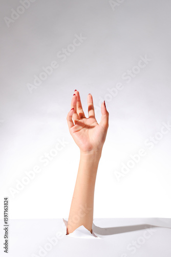 Female hand with red nails and ring rising through torn paper, posed gracefully on white background