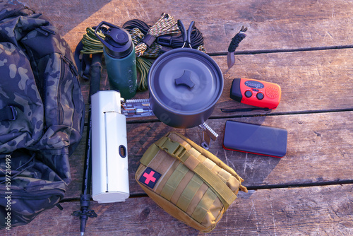 Essential camping gear including backpack, cooking pot, first aid kit, and navigation tools arranged on a weathered wooden surface