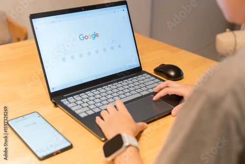 Person using laptop with Google search page open, smartphone and smartwatch on wooden desk, technology concept