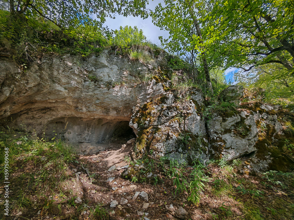 Obraz premium A bright, mossy limestone cliff marks the entrance to the Sima-kő passage cave, surrounded by lush foliage in the Bükk Mountains of Hungary.