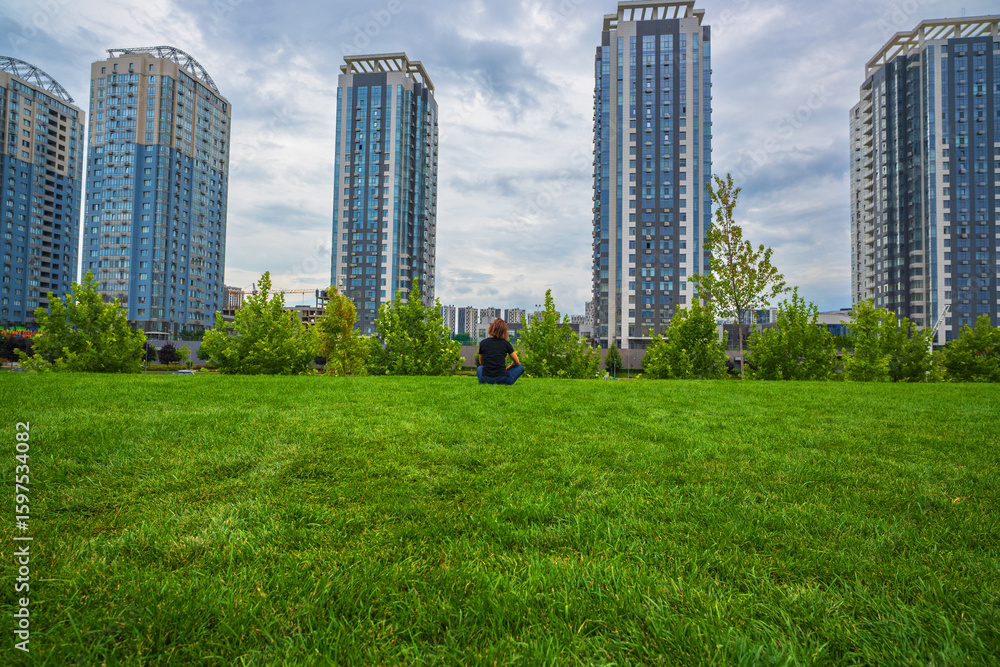 Obraz premium Person relaxing on a vast green lawn, surrounded by modern high-rise buildings under a cloudy sky. A serene blend of urban architecture and nature in the city.
