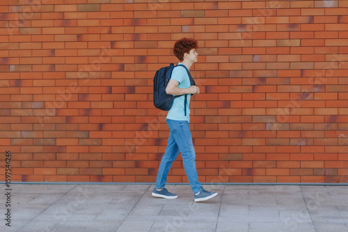 Confident redhead teen walking in casual outfit past urban red brick wall