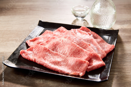 黒毛和牛の霜降りスライス肉を黒い角皿に美しく盛り付け、日本酒の徳利と盃を添えた高級感あふれる和食の食卓シーン。すき焼きやしゃぶしゃぶに最適な素材。