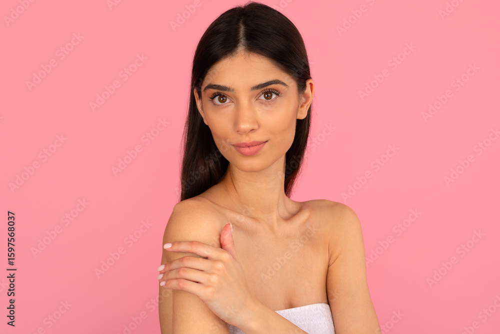 Fototapeta premium Beautiful woman in towel gently holding her shoulder and smiling. Concept of body skincare, health and soft touch on clean pink background, copy space