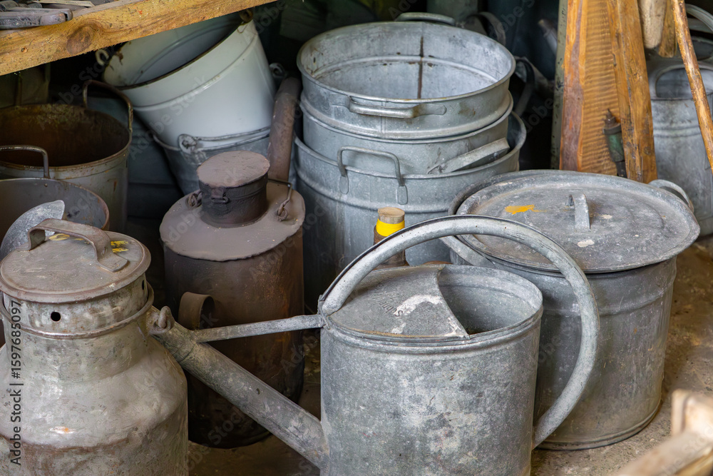 Old metal watering cans, buckets, and milk churns arranged in a rustic storage space. Visible signs of use and patina give the objects a vintage countryside look. Old metal watering cans, buckets, and milk churns arranged in a rustic storage space. Visible signs of use and patina give the objects a vintage countryside look.