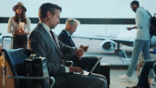 Professional businessman in formal suit working on laptop in busy airport departure lounge. Airplane visible through large window. Corporate travel, remote work, digital nomad lifestyle.