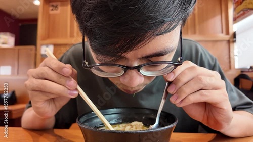 A man with glasses heartily slurps noodles from a bowl of ramen, using chopsticks and a spoon, showcasing the authentic Japanese dining experience.