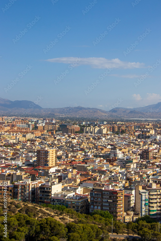 Fototapeta premium Aerial view of Alicante, Spain, showcasing dense urban development with a mix of residential buildings and commercial areas under a clear blue sky. Ideal for cityscapes.