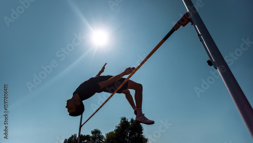 Fototapeta Naklejka Na Ścianę i Meble -  High jumper silhouetted against blue sky during athletic competition in bright sunlight. Concept of training documentation, practice vaults, sports skill refinement