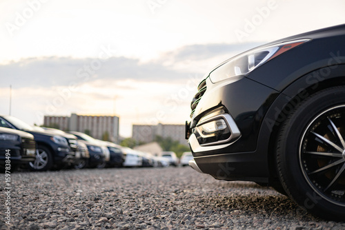 A row of cars parked outside on a gravel lot, featuring a close-up of a black vehicle's front.
