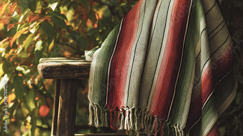 A traditional embroidered serape poncho in bold stripes of red, green, and white, draped over a rustic wooden chair with soft sunlight.