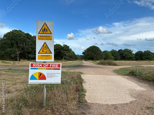 Warning signs reading 'Troops Training' and 'Risk of Fire' alert posted along woodland paths. Rural military training area natural setting with blue sky on summers day