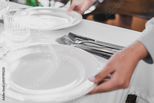 Fotografie Close-up of elegant table setting with person placing a white plate on a white tablecloth, featuring glassware, cutlery, and napkins in a formal dining environment