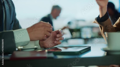 A business meeting is in progress, with a tablet and coffee cup on the table. The setting seems to be a modern office or cafe.