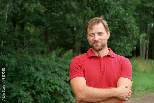 Portrait of handsome young man in a red t-shirt outdoors. Male with fair hair and beard feel happy, calm and serious. Green nature background. Guy looking away into distance. Diversity People