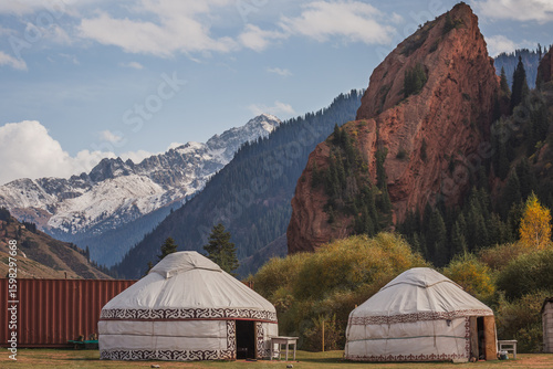 Two traditional yurts stand in the scenic Charyn Canyon, Kazakhstan, nestled amongst vibrant autumn foliage and majestic mountains. Breathtaking landscape of snow-capped peaks and red rock formations.