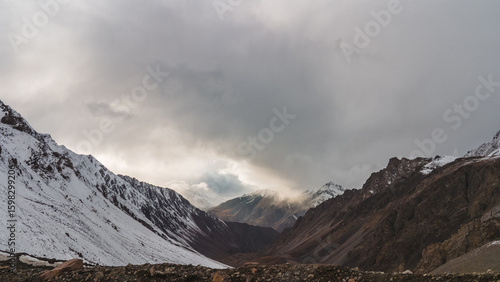 Dramatic panoramic view of snow-capped mountains under a cloudy sky.  The rugged terrain and the interplay of light and shadow create a breathtaking scene.  A powerful image of nature's grandeur.