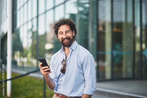A man in a light blue shirt and sunglasses stands outside a sleek office building, smiling as he uses his smartphone in the warm afternoon light.