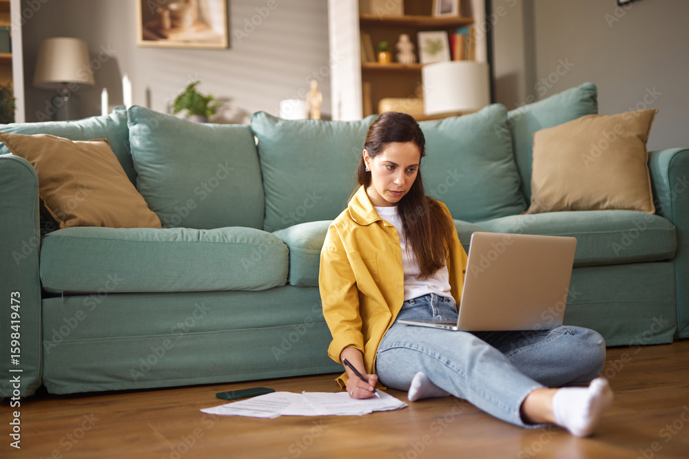 Naklejka premium A person is working intently on a laptop while seated on the floor, surrounded by a cozy living room with a comfortable couch and scattered documents, enjoying a productive moment.