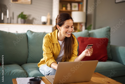 A woman is sitting comfortably on a couch in her living room, joyfully shopping online using a laptop. She holds a credit card, looking pleased while browsing.